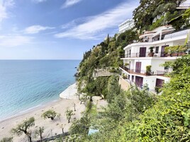 A coastal hotel with balconies overlooking the sea, a beach, and lush greenery.