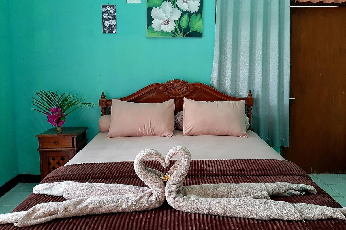 A neatly made bed with a heart-shaped towel arrangement, a wooden headboard, and a teal wall with floral artwork.