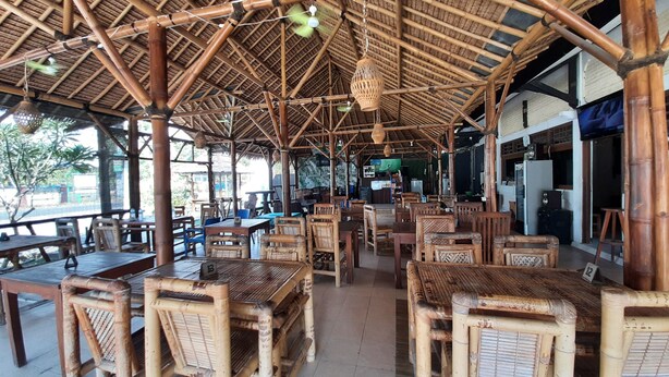 A bamboo-roofed restaurant with wooden tables and chairs, a TV screen, and a visible outdoor area. - New Taman Sari Homestay (Pemenang)