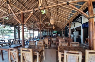 Un restaurante con techo de bambú, mesas y sillas de madera, una pantalla de televisión y un área exterior visible.