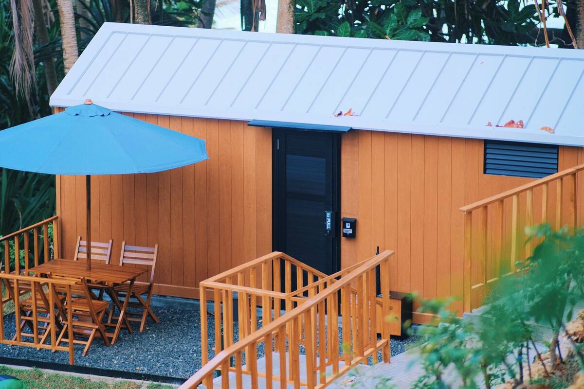 Uma cabana de madeira com varanda, mesa e cadeiras, e um guarda-sol azul.