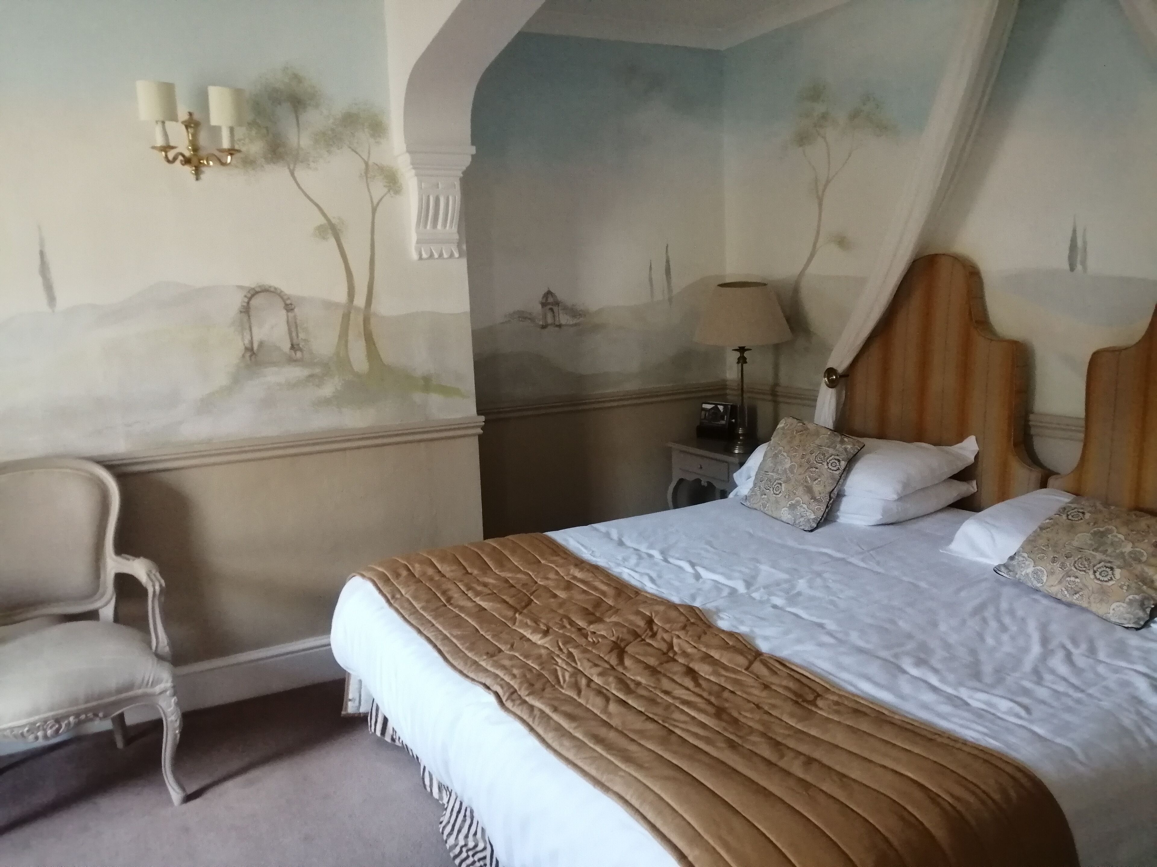 Une chambre à coucher avec un grand lit, une table de chevet, une lampe, une chaise et une fresque murale.