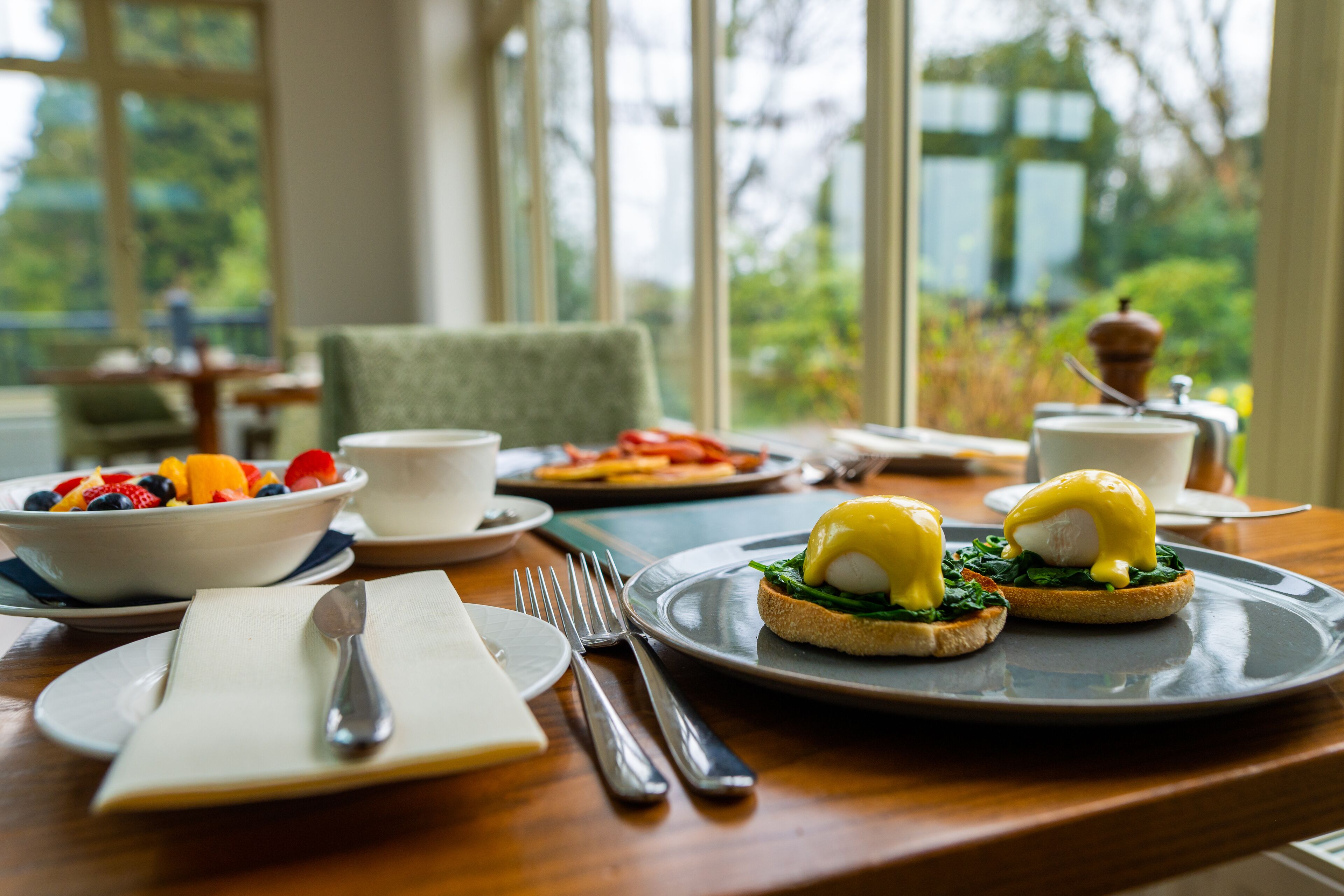 A breakfast setting with eggs benedict on toast, a bowl of fruit, and a cup of coffee on a wooden table with a view of greenery outside.