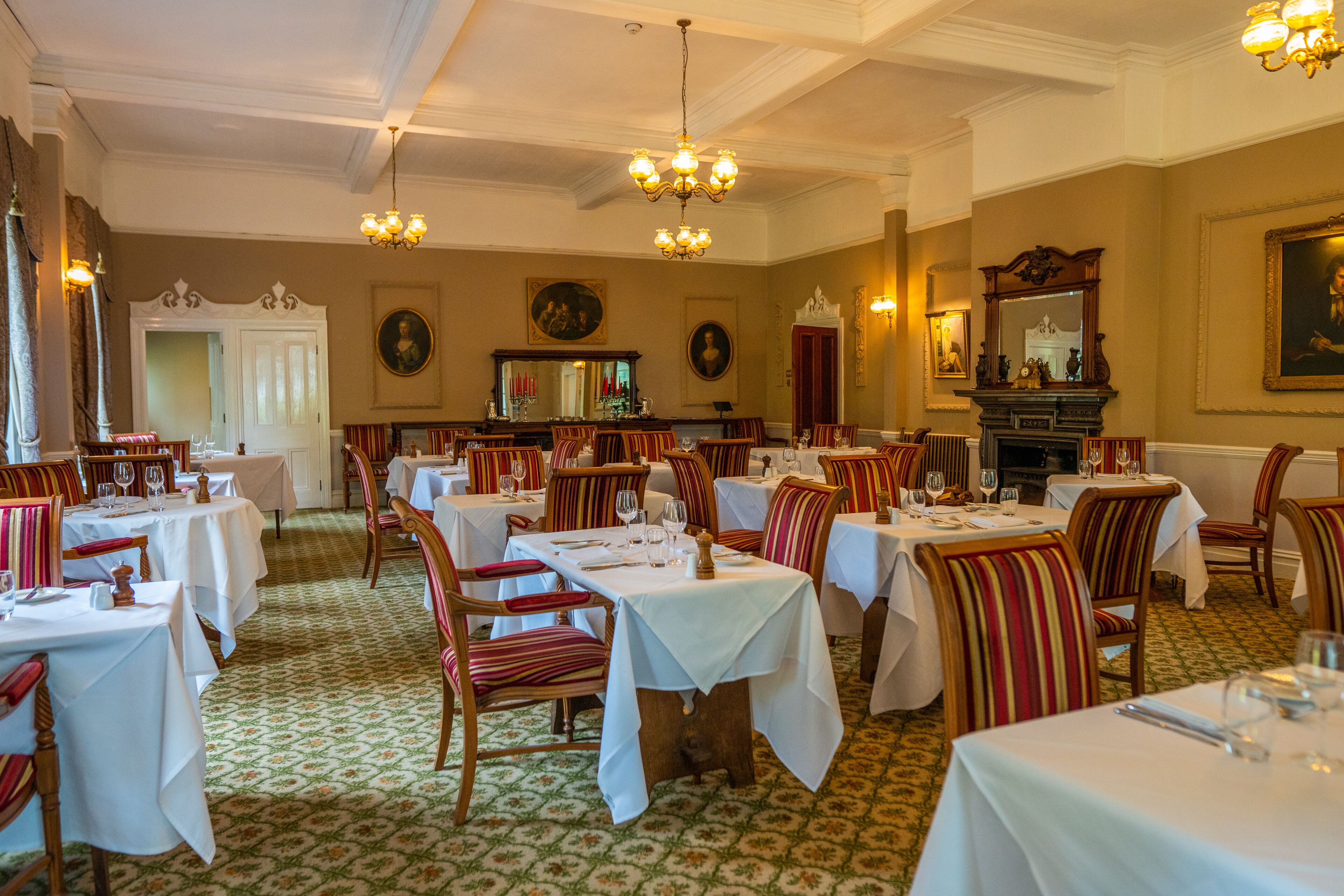 A formal dining room with multiple tables and chairs, chandeliers, and wall-mounted paintings.