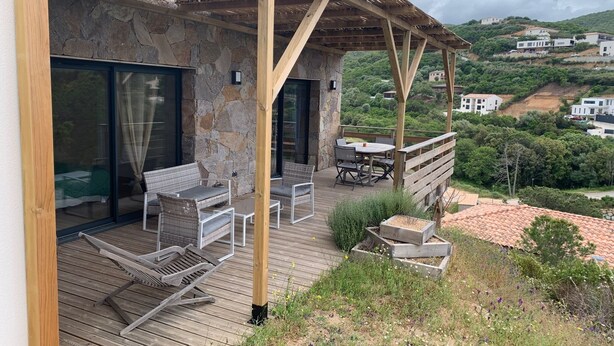 A wooden deck with outdoor furniture, a stone wall, and a view of a hillside with houses. - Jolie Dépendance Avec sa Très Belle vue mer (Pietrosella)