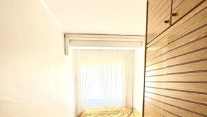 Une petite pièce étroite, avec un lit, une armoire en bois et une fenêtre ornée de rideaux légers.