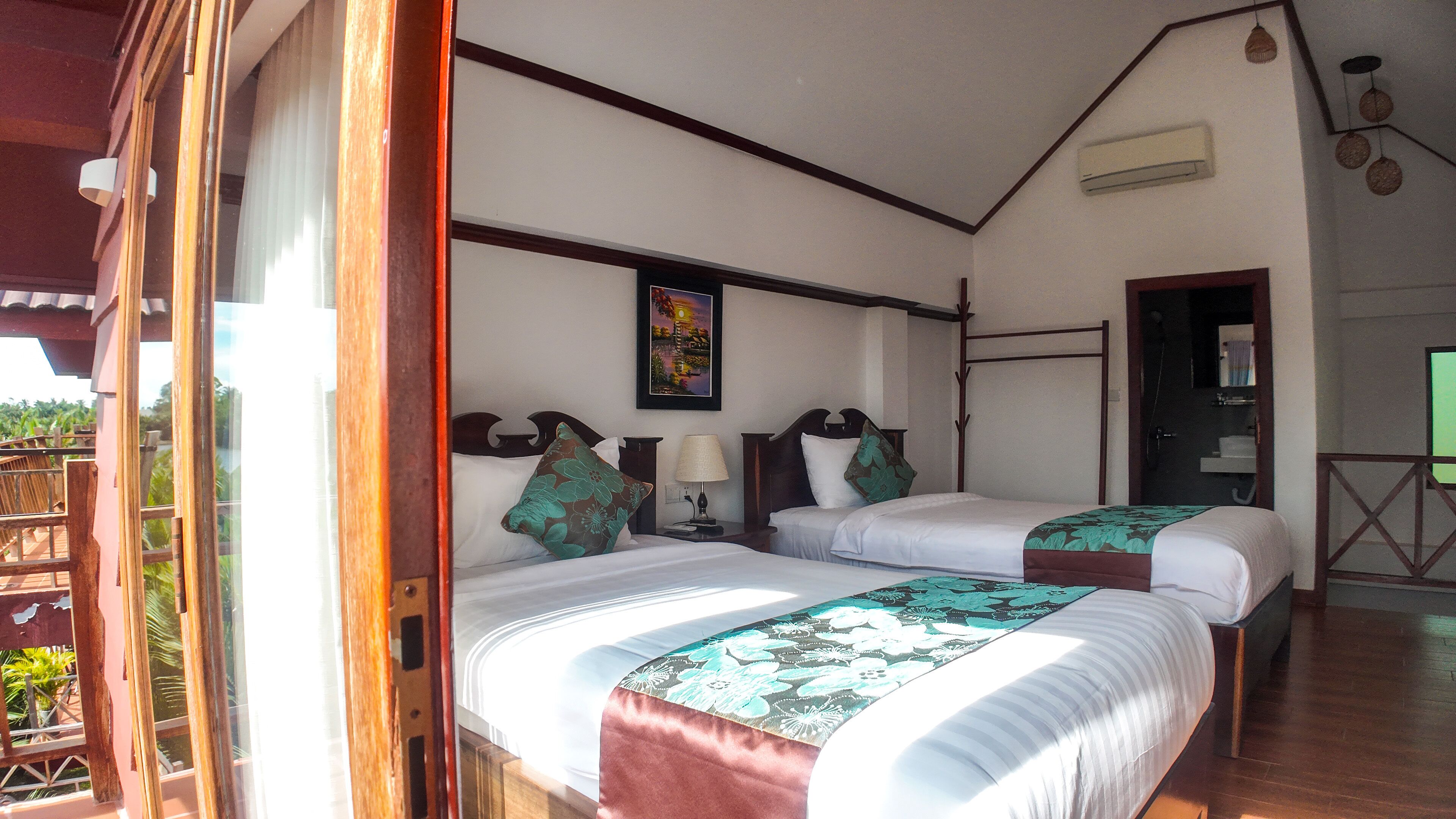 A room with two beds, a painting on the wall, a lamp, and a view of the outdoors.