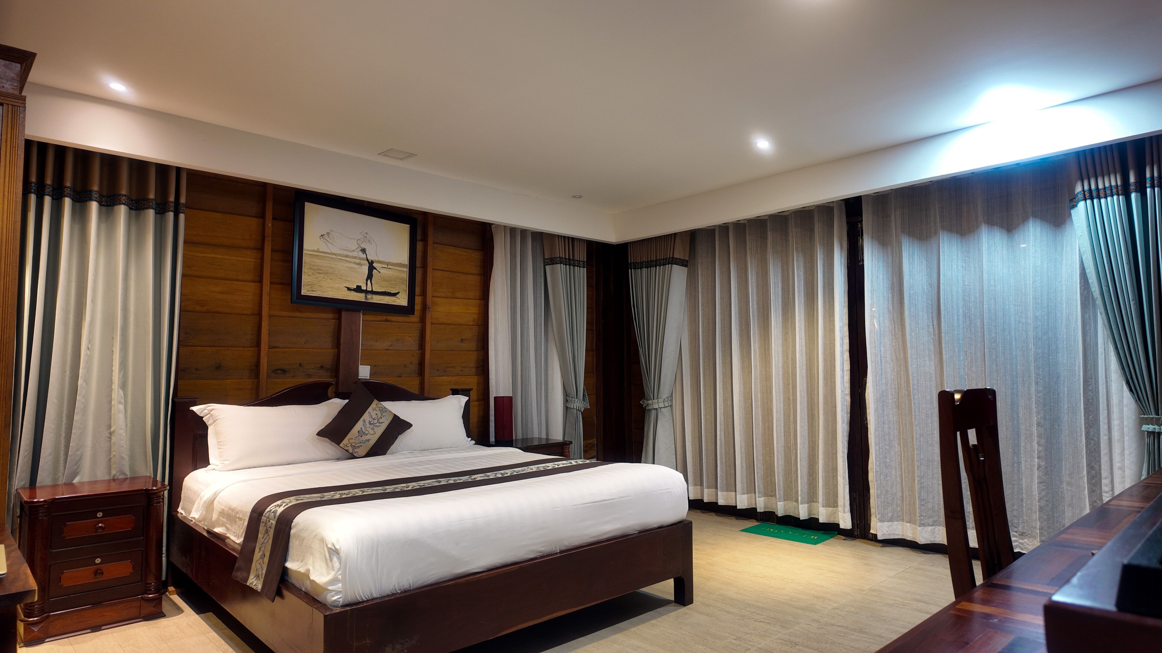 Ein Hotelzimmer mit einem großen Bett, einem Holzkopfteil und einem Bild von einer Person beim Angeln. Es gibt einen Schreibtisch mit einem Stuhl, einen Nachttisch und eine grüne Matte auf dem Boden.
