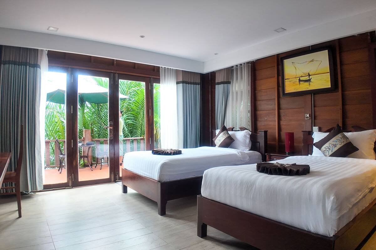 Habitación de hotel con dos camas, piso de madera, balcón con mesa y sillas, y un ventanal grande con cortinas.