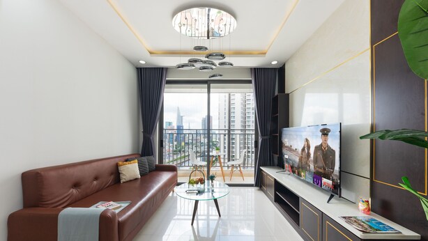 A modern living room with a brown leather sofa, a glass coffee table, a flat-screen TV, and a city view through large windows. - RIVERGATE apartment infinity pool & Gym  (Ho Chi Minh City)