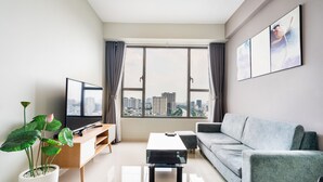 A modern living room with a large window, a flat-screen TV, a grey sofa, a coffee table, and a potted plant. - RIVERGATE apartment infinity pool & Gym  (Ho Chi Minh City)