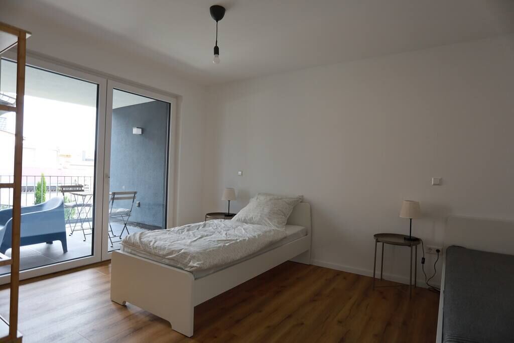A bedroom with a single bed, a small bedside table, a chair, and a balcony with a table and chairs.
