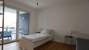 A bedroom with a single bed, a small bedside table, a chair, and a balcony with a table and chairs.