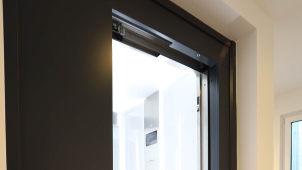 A modern elevator with a dark exterior and a glass door, leading to a well-lit interior space.