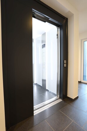 Ein moderner Aufzug mit dunkler Außenverkleidung und einer Glastür, der in einen gut beleuchteten Innenraum führt.