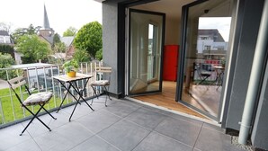 A balcony with a table and chairs, a sliding glass door, and a view of a church and greenery.