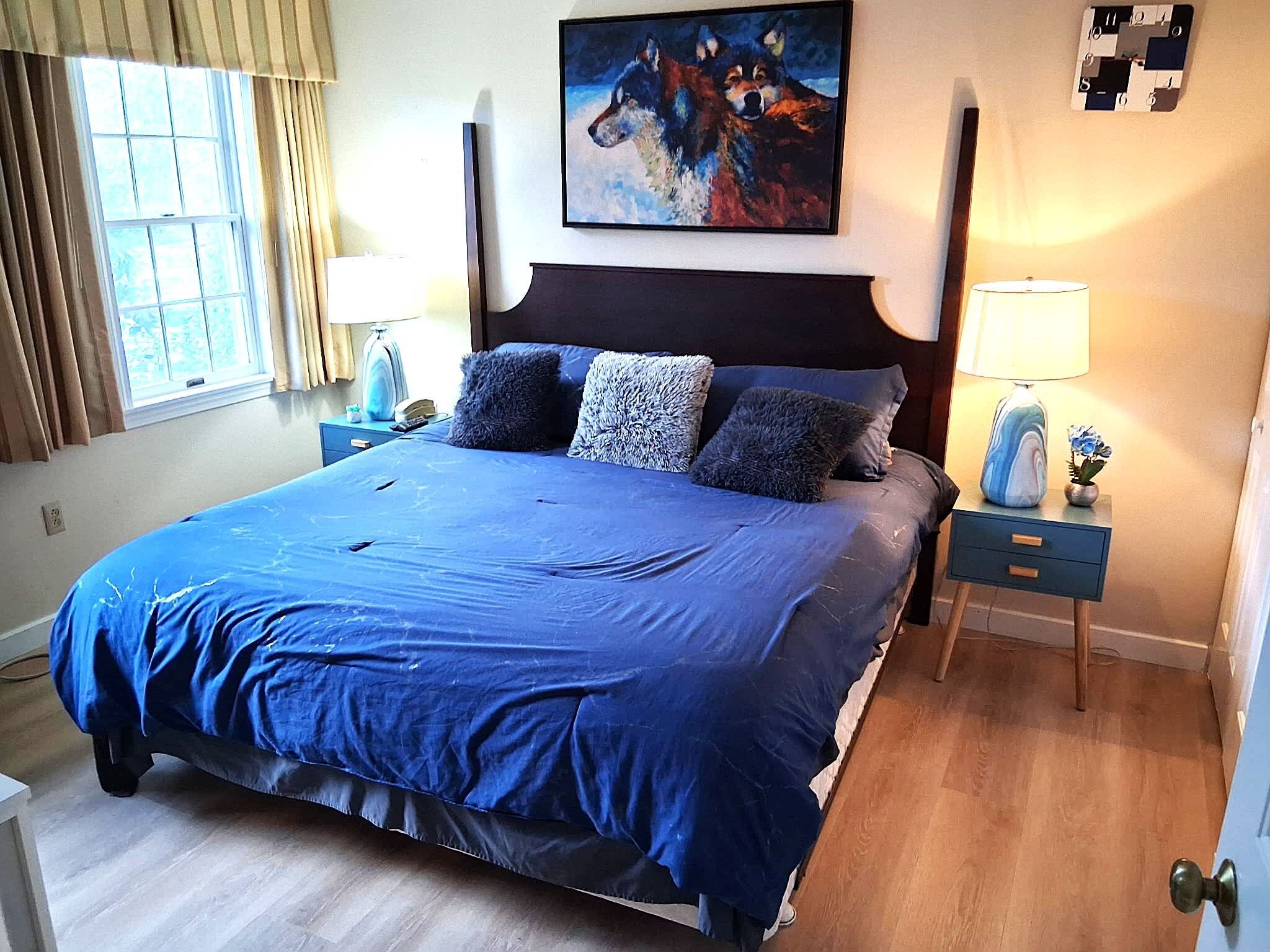 A hotel room with a large bed, a painting on the wall, a nightstand with a lamp, and a window with curtains.