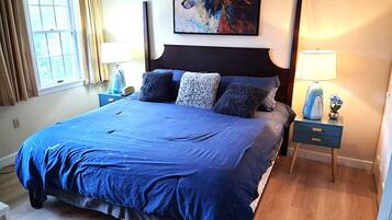 A hotel room with a large bed, a painting on the wall, a nightstand with a lamp, and a window with curtains.