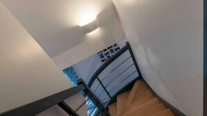 Una escalera con peldaños de madera y barandilla de metal, que conduce a una plataforma con pared blanca y una lámpara en el techo.