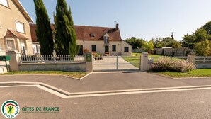 Une entrée fermée par un portail mène à un bâtiment doté d’un toit en tuiles, entouré d’une clôture blanche et de verdure.