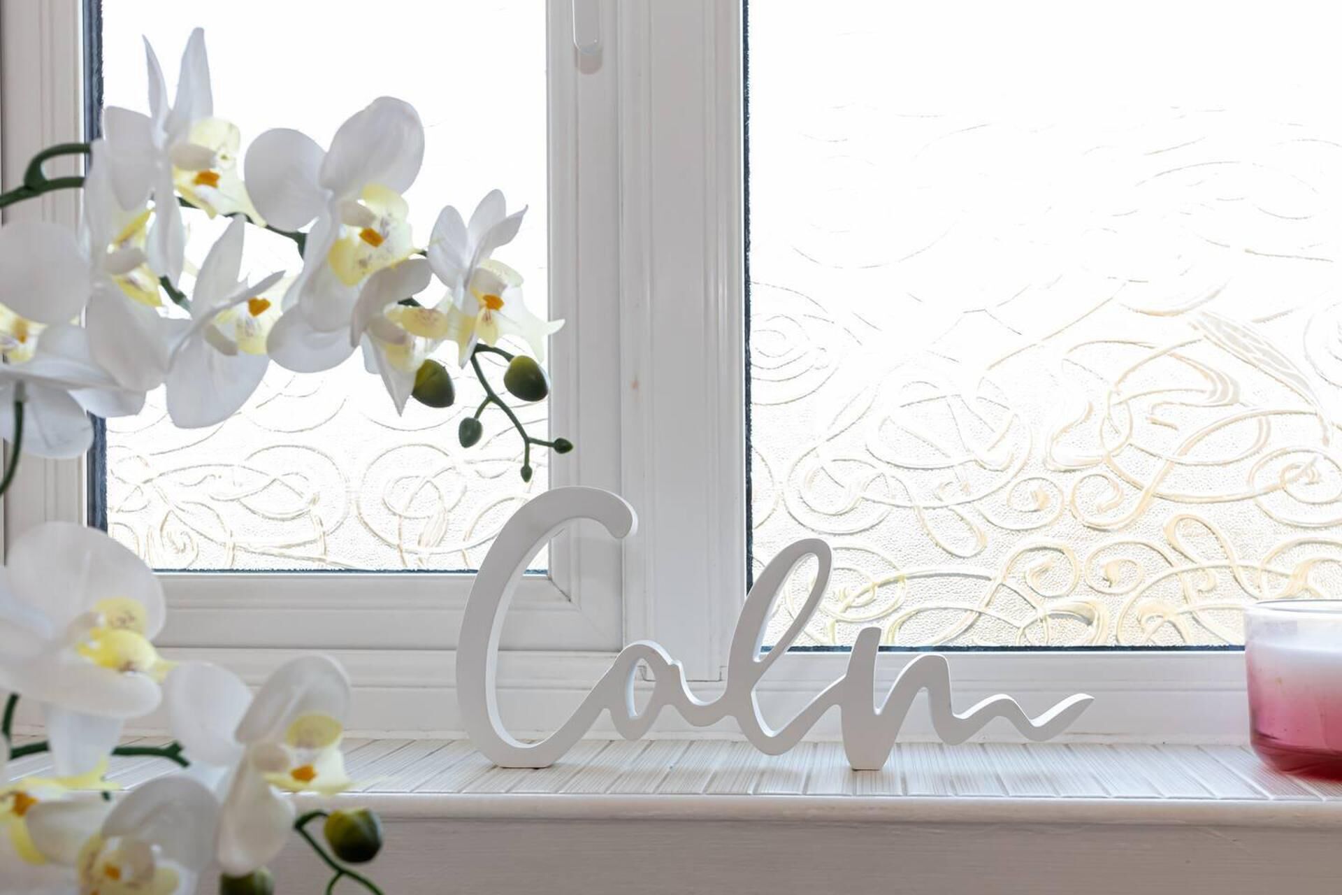 A window with a frosted glass pattern, a white decorative sign spelling "Calm," and white orchids on the window sill.