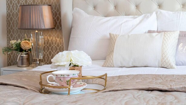 Una cama bien hecha con un cabecero capitonado, una lámpara de noche, un jarrón con flores y una bandeja con una taza y platillo de té.