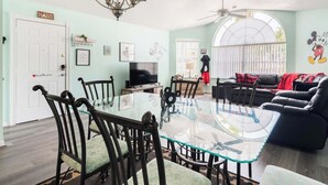 A spacious living room with a glass-top dining table surrounded by chairs, a black leather sofa, and a ceiling fan. - Disney, Pool, Hot Tub, Gym! (Kissimmee)