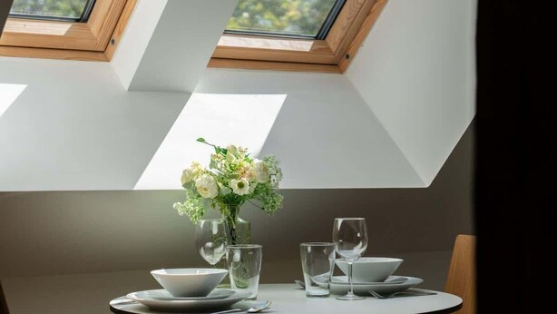 A dining table with a vase of flowers, clear glassware, and plates, set in a room with skylights. - Tasteful Apartment in Poznan | Private Parking (Poznan)