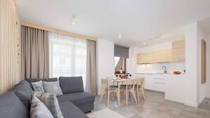 A modern living room with a grey sofa, a dining area with a table and chairs, and a kitchen with white cabinets and a wooden countertop.