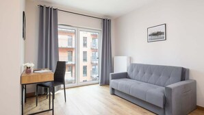 Un salon moderne avec un canapé gris, un bureau en bois avec une chaise et une grande fenêtre agrémentée de rideaux gris.