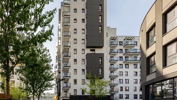 Un moderno edificio de apartamentos con varios balcones, un parque arbolado y un local comercial visible.