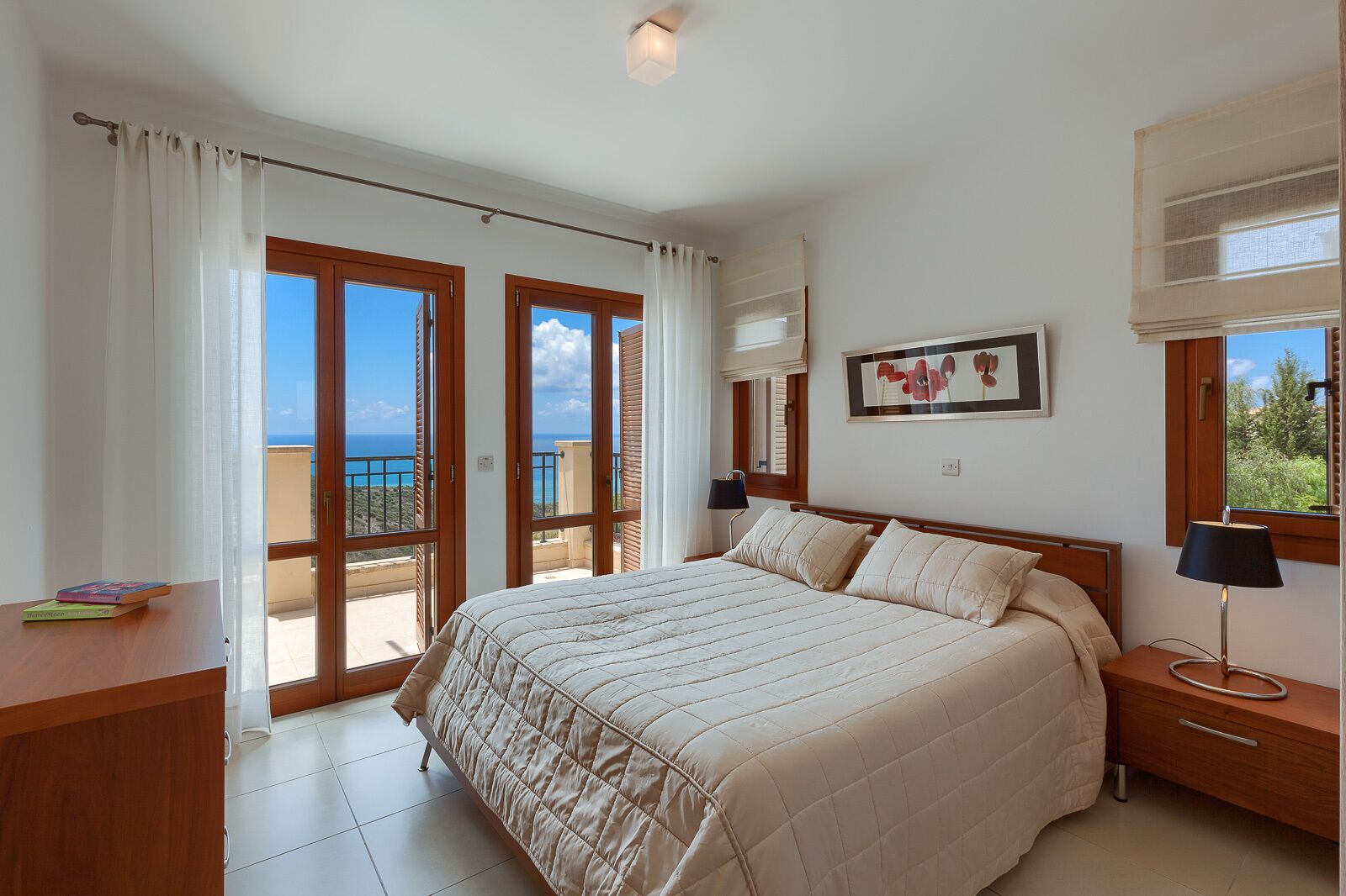 A bedroom with a bed, a nightstand, a desk, and a view of the sea through large windows.