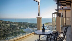 A balcony with a table, chairs, a bottle of wine, and a glass. The balcony overlooks a scenic view of the sea and hills.