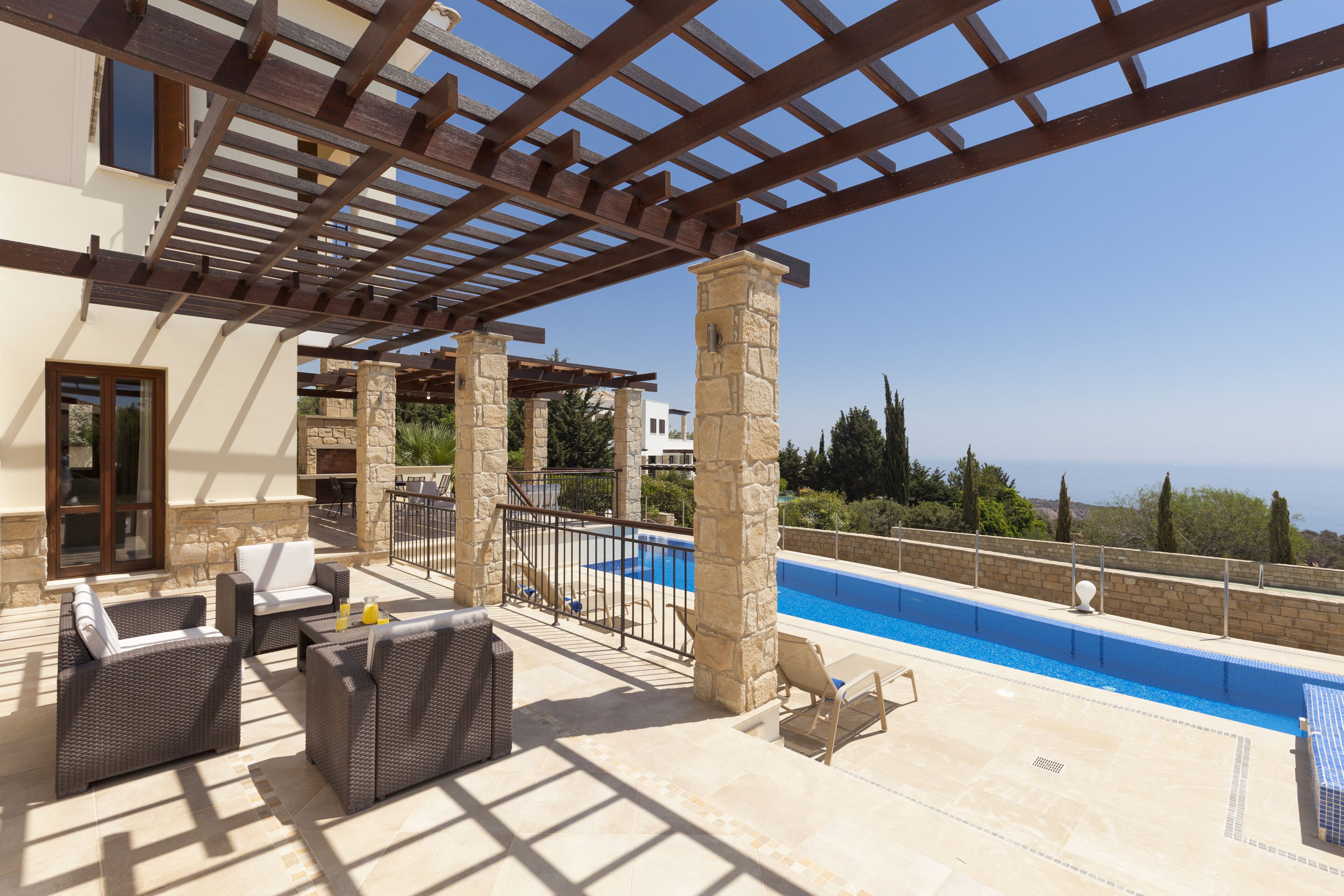 Un área junto a la piscina con mobiliario de mimbre, pilares de piedra y una pérgola.