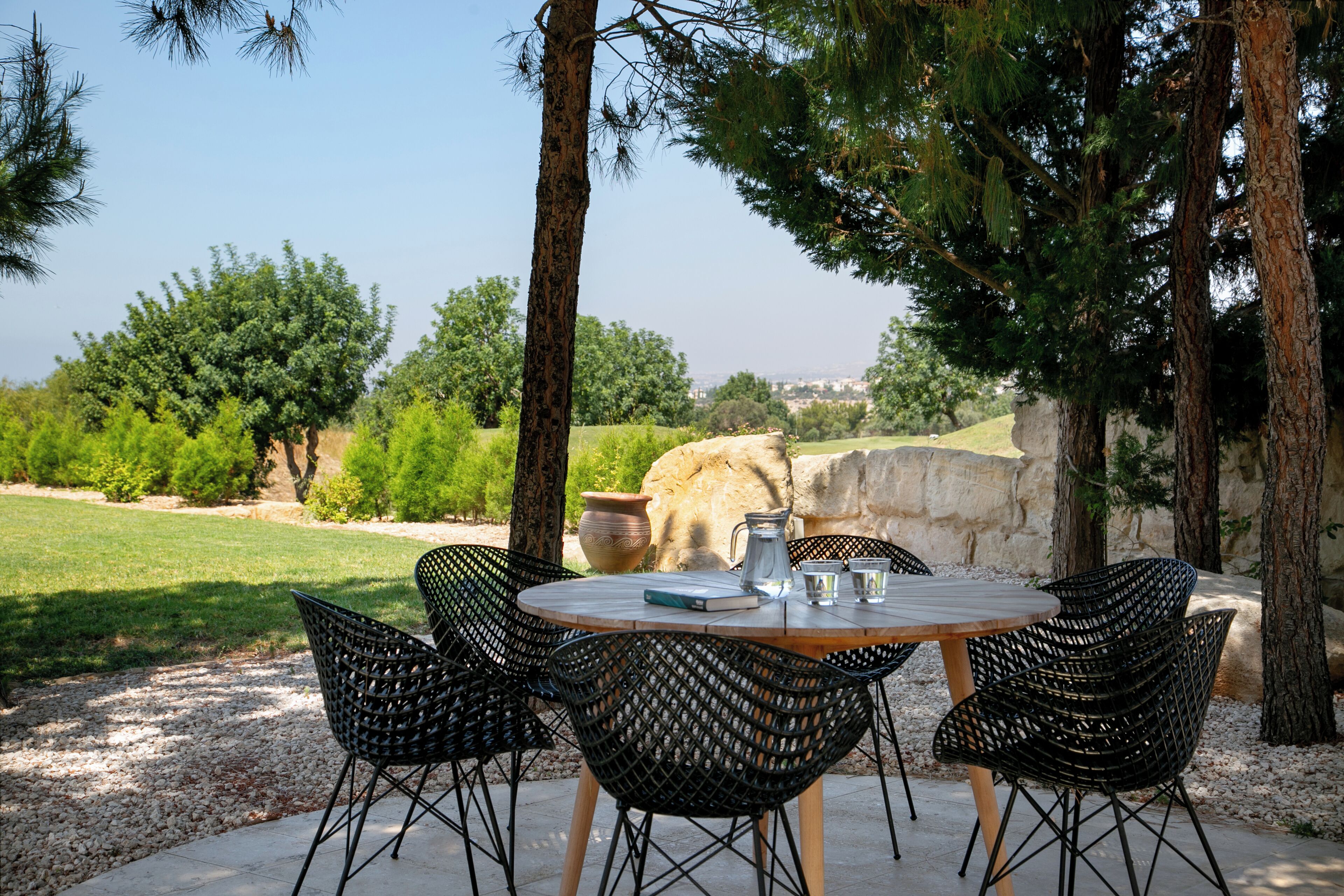 A round wooden table with four black woven chairs, surrounded by trees and a stone wall, set on a gravel path.