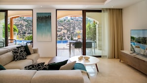 A modern living room with a sofa, coffee table, and TV. It features large windows with a view of a pool and hills.