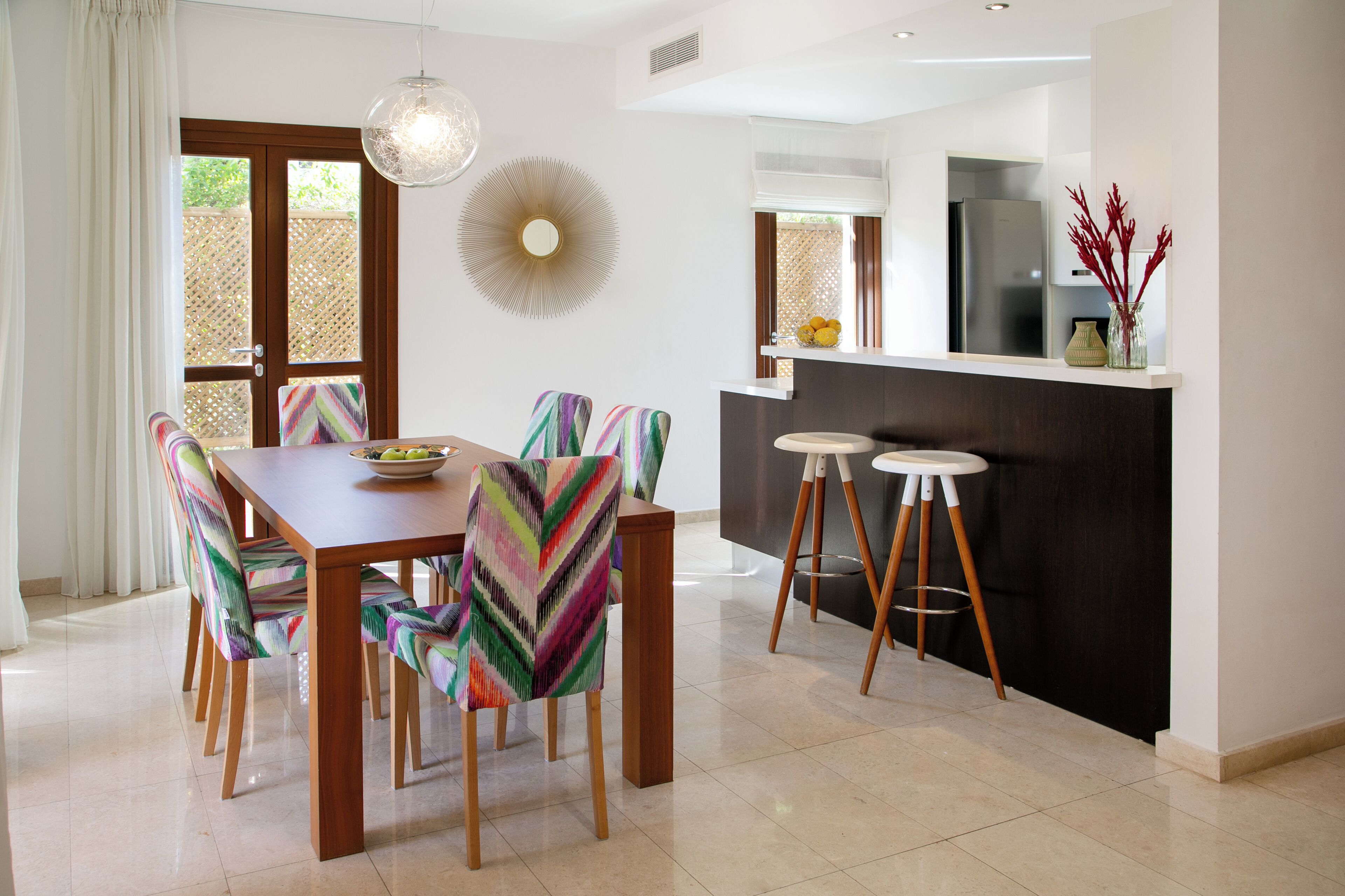 Un comedor con mesa de madera y sillas coloridas, una lámpara colgante moderna y una encimera de cocina con taburetes.