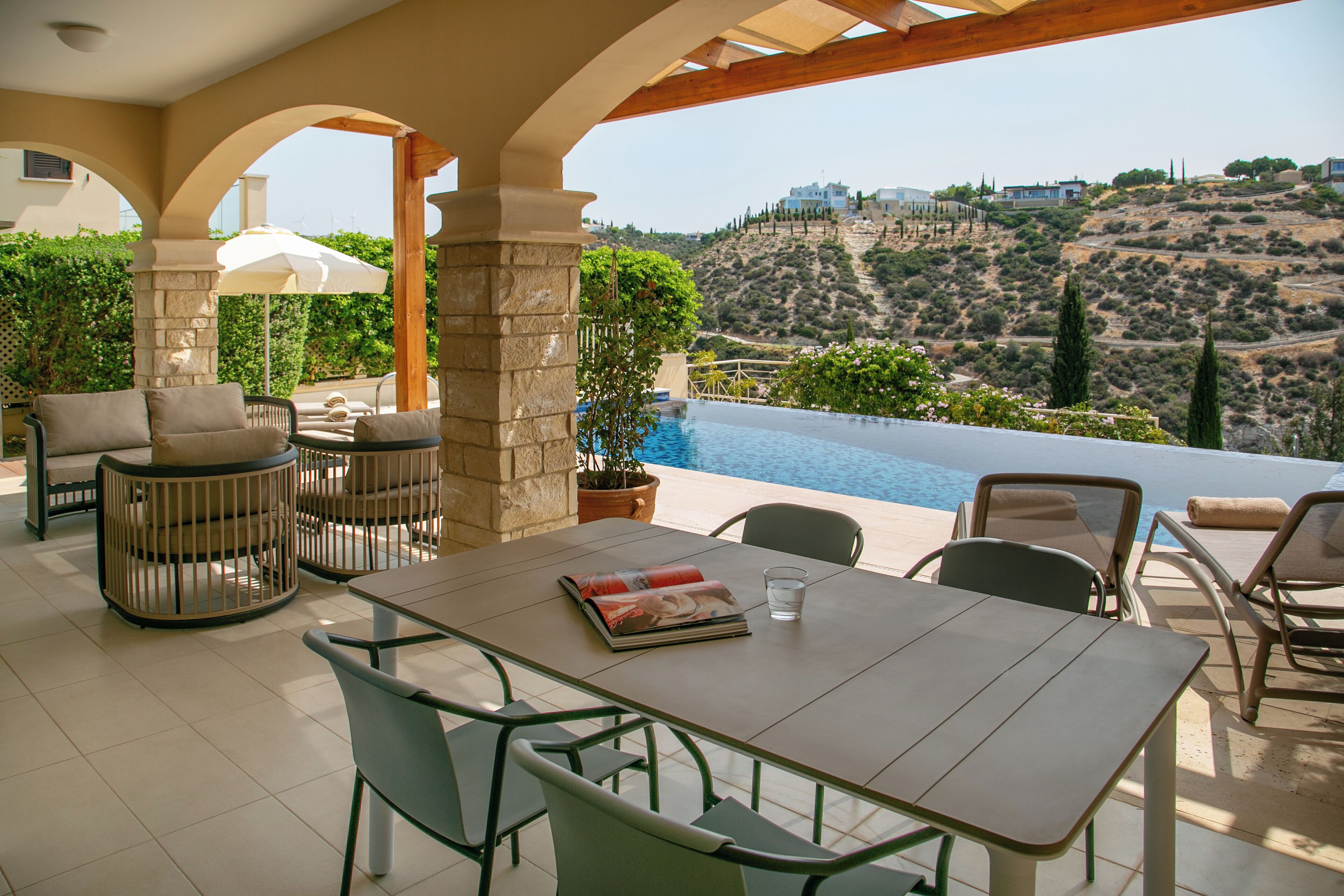Un espacio al aire libre cubierto con vistas a la piscina, pilares de piedra y una mesa con revistas y un vaso.