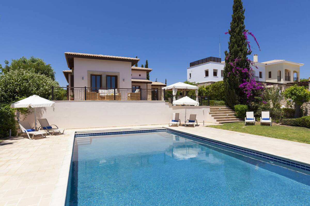 A swimming pool with lounge chairs and umbrellas, surrounded by a landscaped garden and two buildings.