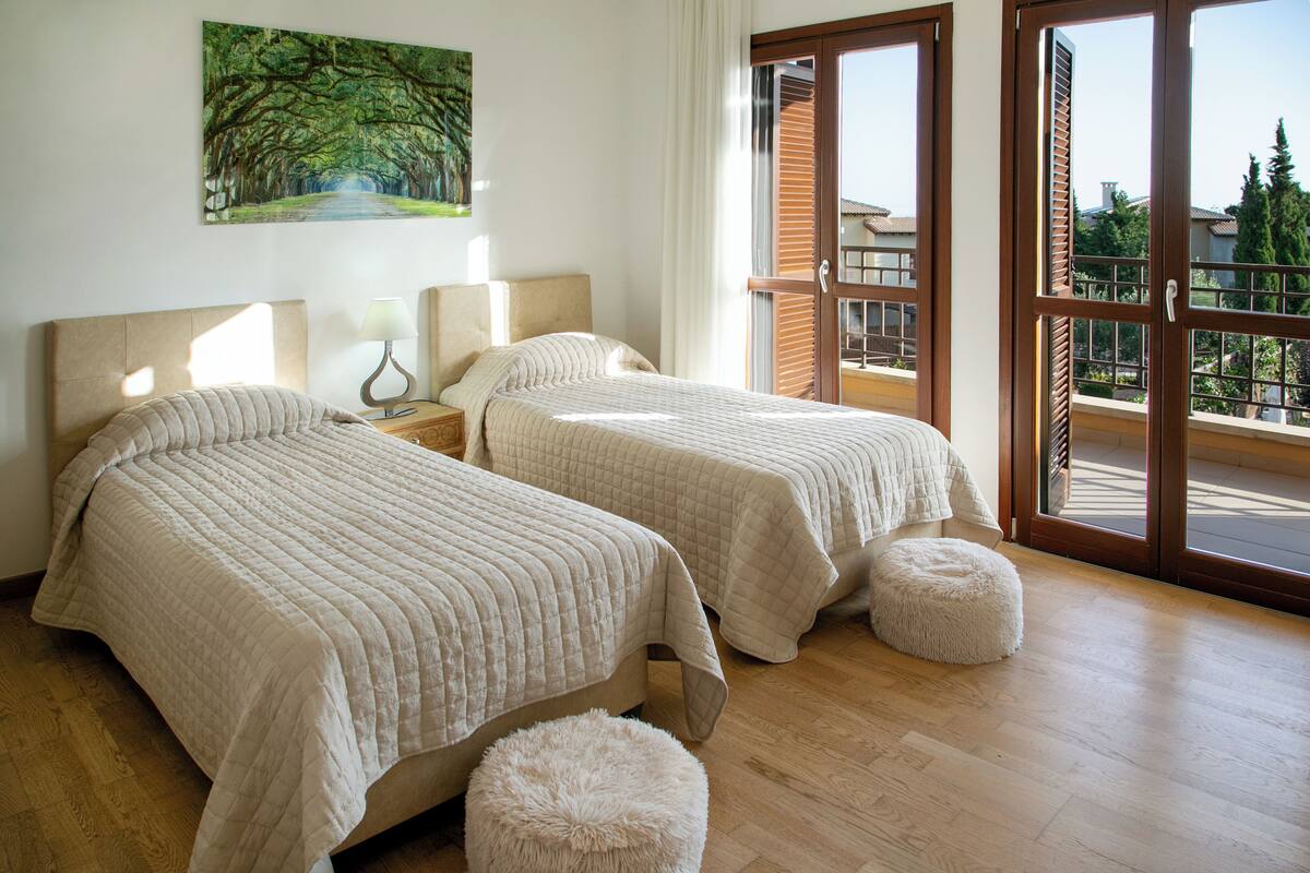 A hotel room with two beds, a painting on the wall, and a balcony with a view.