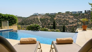 A pool with lounge chairs and towels, overlooking a hilly landscape with buildings.
