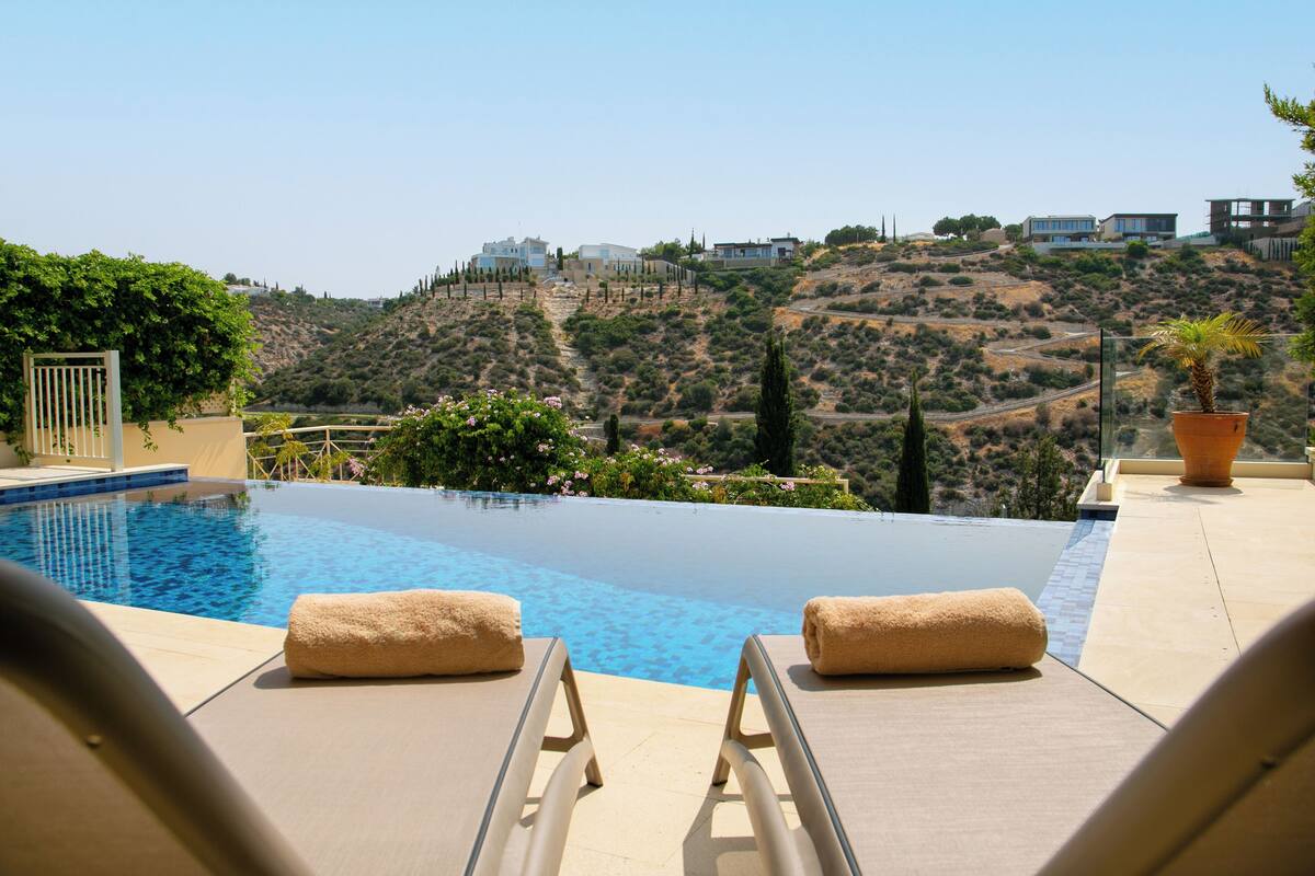 A pool with lounge chairs and towels, overlooking a hilly landscape with buildings.