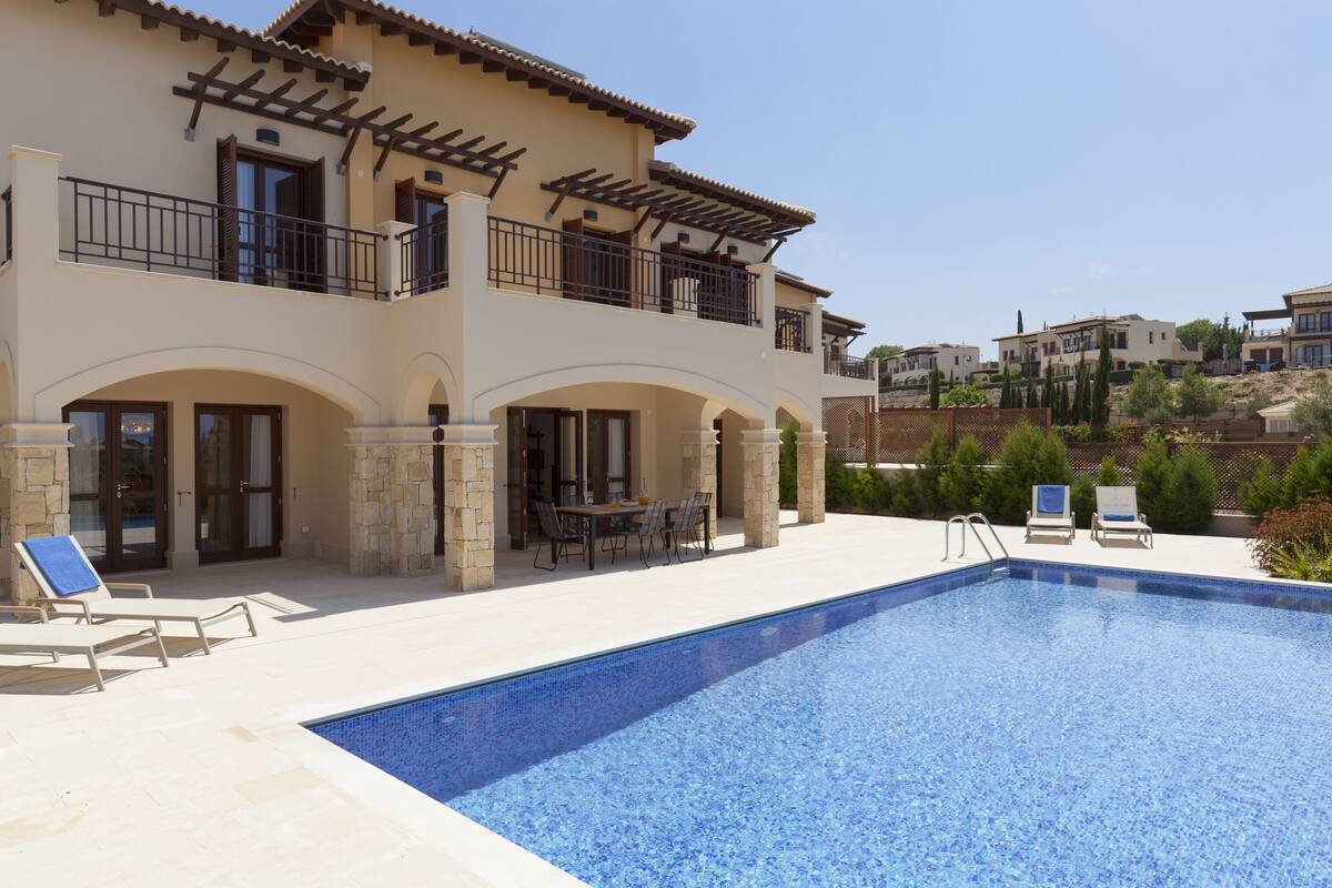 A pool area with lounge chairs and a dining table, surrounded by a building with balconies and a view of other houses.