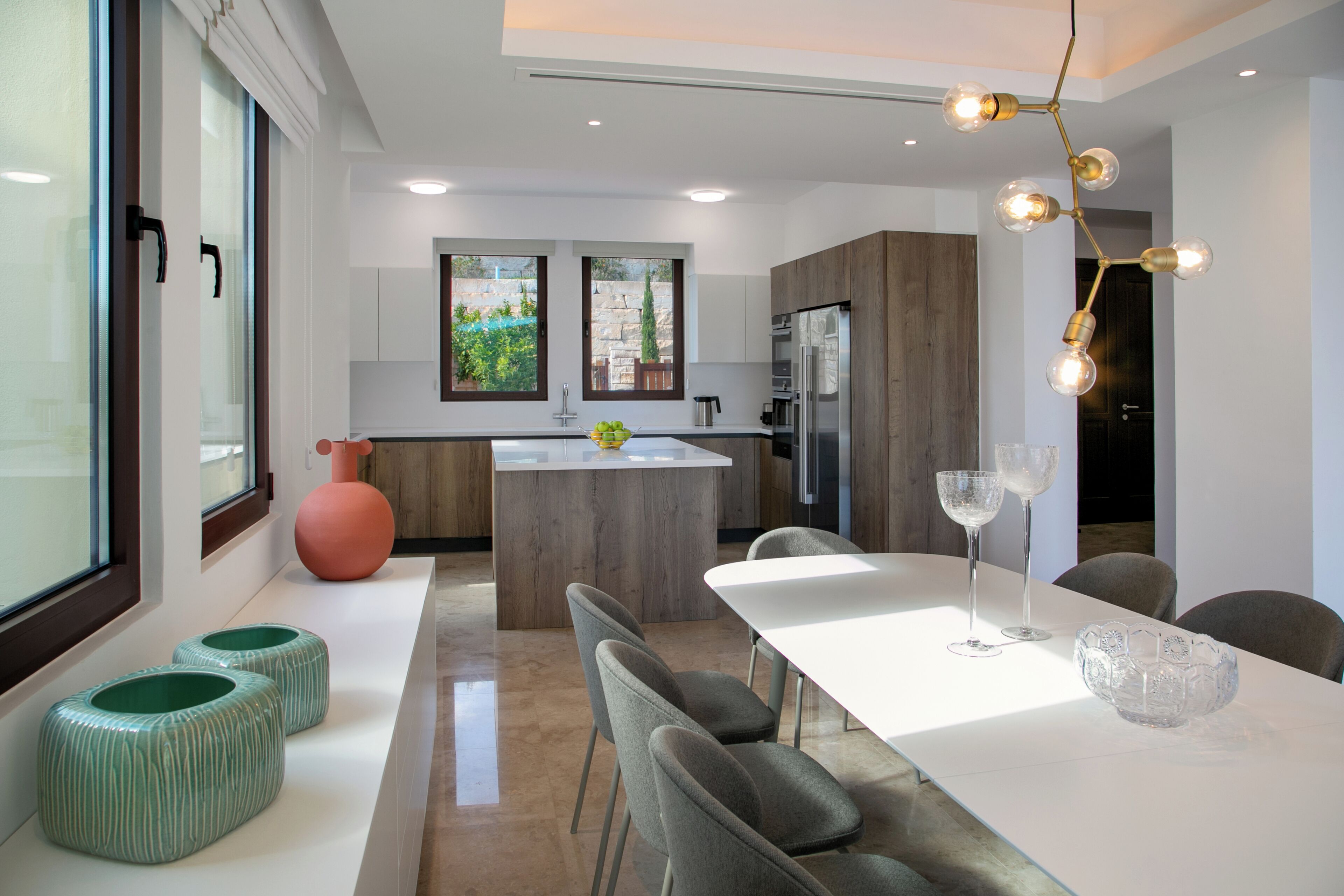 Una cocina moderna con una isla central, un área de comedor con una mesa blanca y un ventanal que ofrece vistas a zonas verdes.