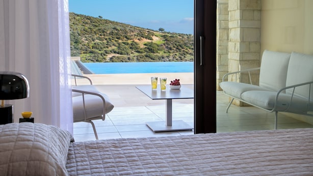 A bedroom with a bed, a bedside table with a lamp, and a view of a balcony with a table and chairs overlooking a beach and ocean.