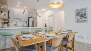 A modern kitchen with white cabinets, a dining area with a wooden table and chairs, and a kitchen island with a refrigerator and sink. - Le Refuge - Revelstoke Condo with Hot Tub (Revelstoke)