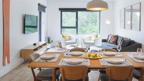 A dining area with a wooden table set for four, surrounded by chairs, with a large window providing a view of greenery. - Le Refuge - Revelstoke Condo with Hot Tub (Revelstoke)
