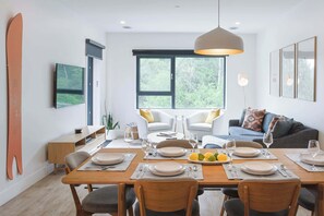 A dining area with a wooden table set for four, surrounded by chairs, with a large window providing a view of greenery.