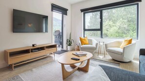 A modern living room with a flat-screen TV, a wooden entertainment unit, a round coffee table, two white armchairs with yellow cushions, and a large window with a view of greenery. - Le Refuge - Revelstoke Condo with Hot Tub (Revelstoke)