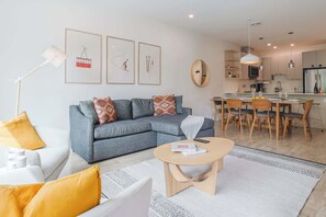 Een moderne woonkamer met een grijze bank, een salontafel, en een eethoek met houten stoelen, met een keuken op de achtergrond.